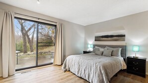 Un dormitorio con un amplio ventanal corredera que da a un área exterior, una cama con cabecera gris, dos mesitas de noche con lámparas y una obra de arte colgada en la pared.