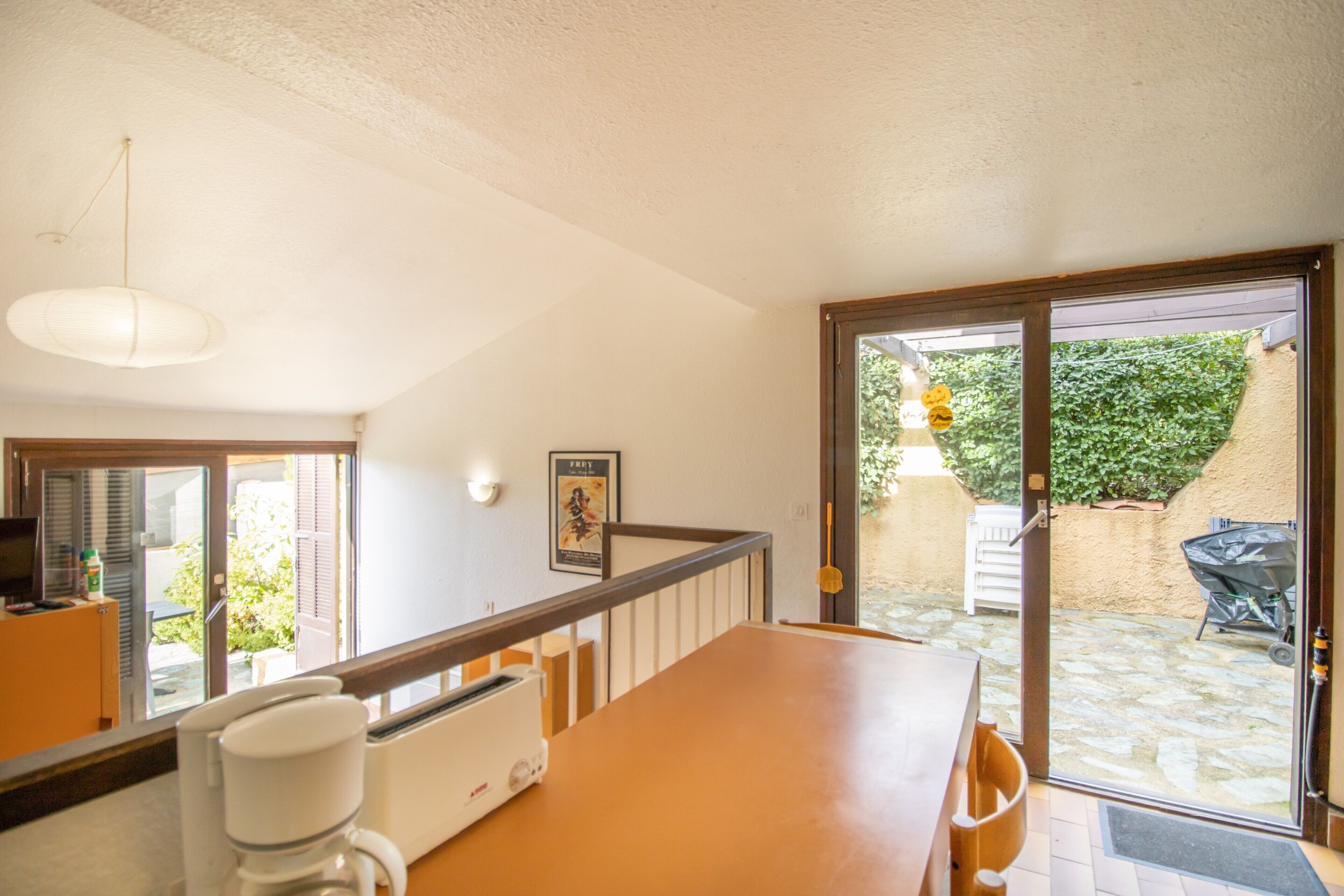 A dining area with a wooden table and chairs, a coffee machine, and a view of a garden through a glass door.