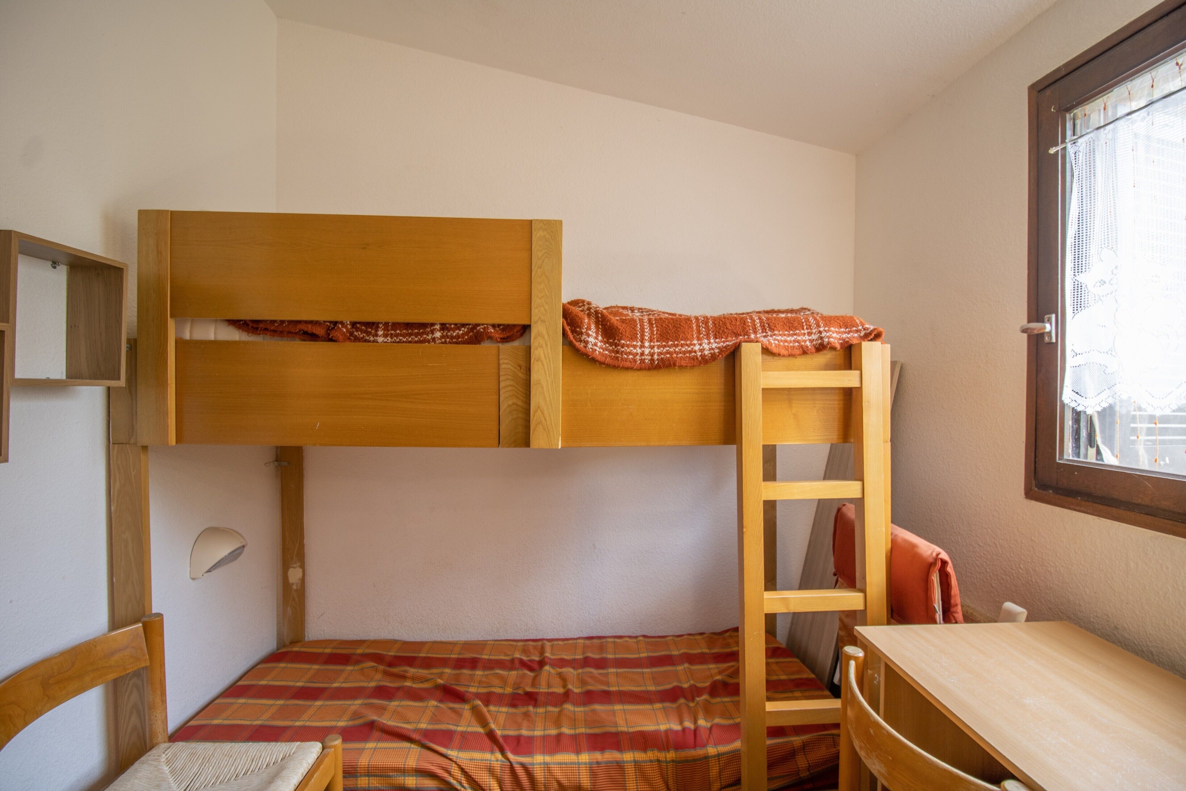 A bunk bed with a lower bed and a desk, a ladder, and a window with lace curtains.
