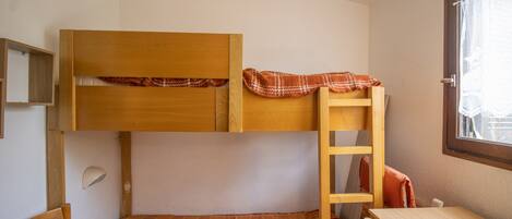 A bunk bed with a lower bed and a desk, a ladder, and a window with lace curtains.