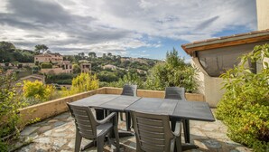 Eine Terrasse im Freien mit Tisch und Stühlen, umgeben von Grünpflanzen und mit Blick auf ein Hügeldorf.