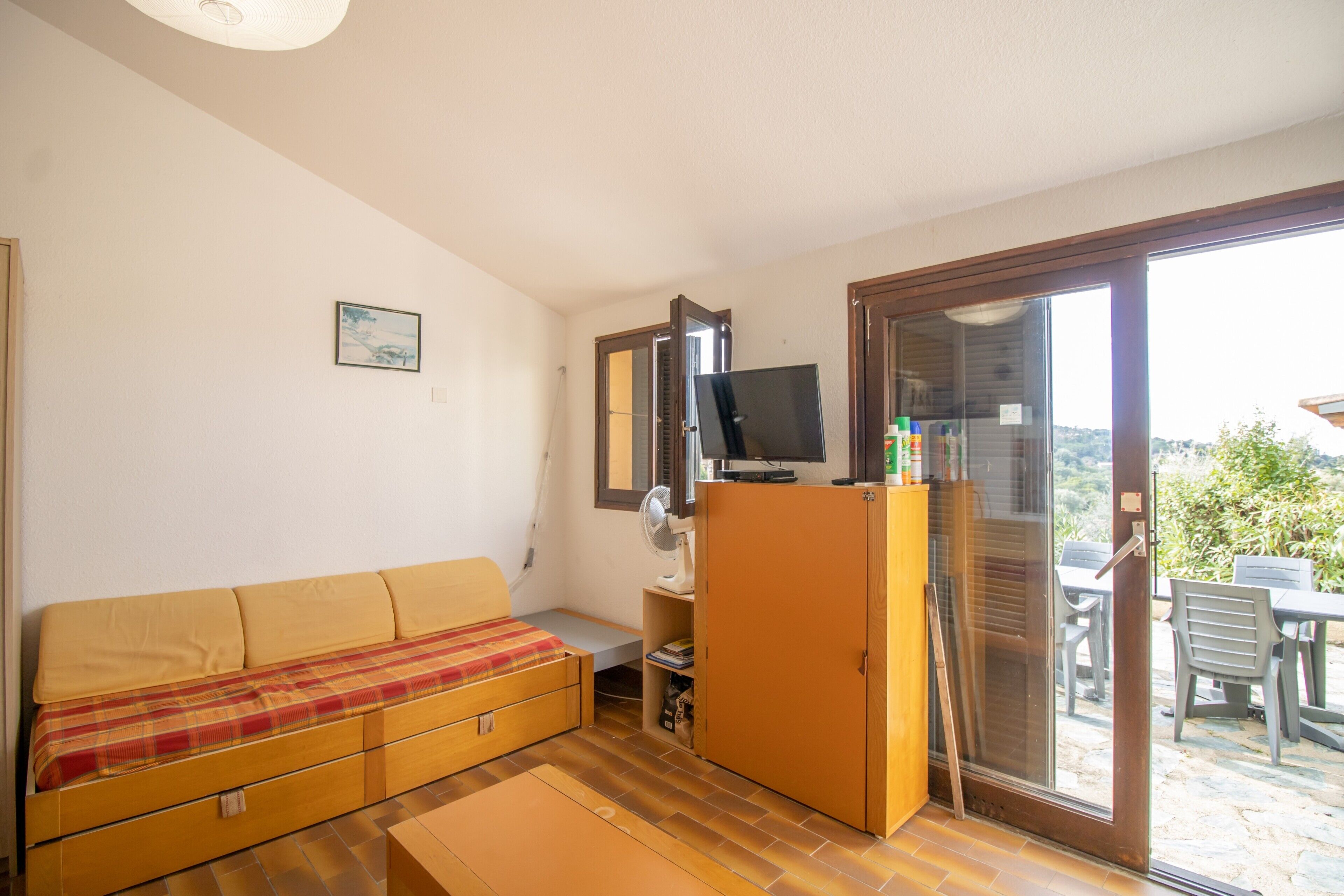 Une pièce comprenant un canapé, un téléviseur posé sur un meuble, un placard et une porte-fenêtre coulissante donnant sur un espace extérieur avec une table et des chaises.
