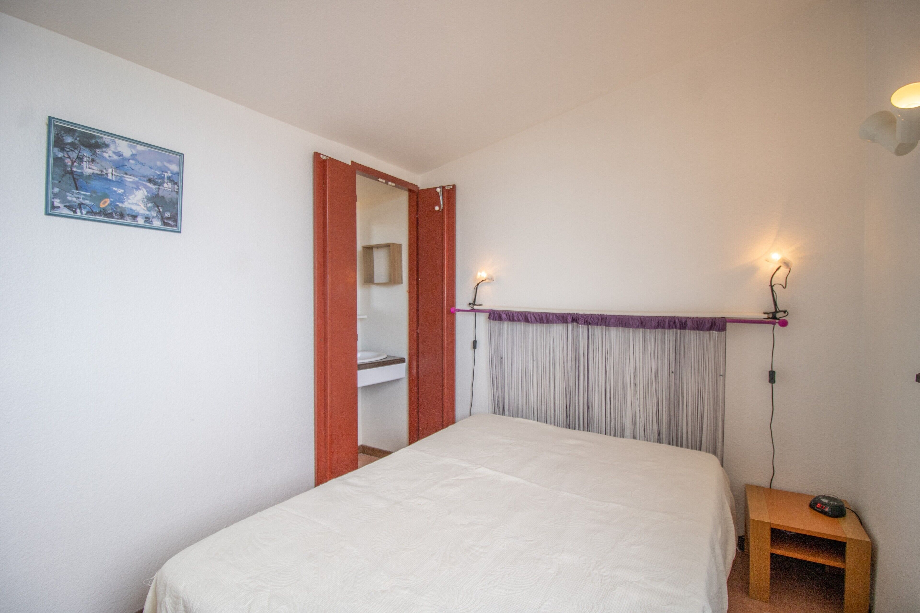 Una camera da letto con un letto, un comodino con una sveglia, un quadro appeso al muro e una porta che dà su un bagno.