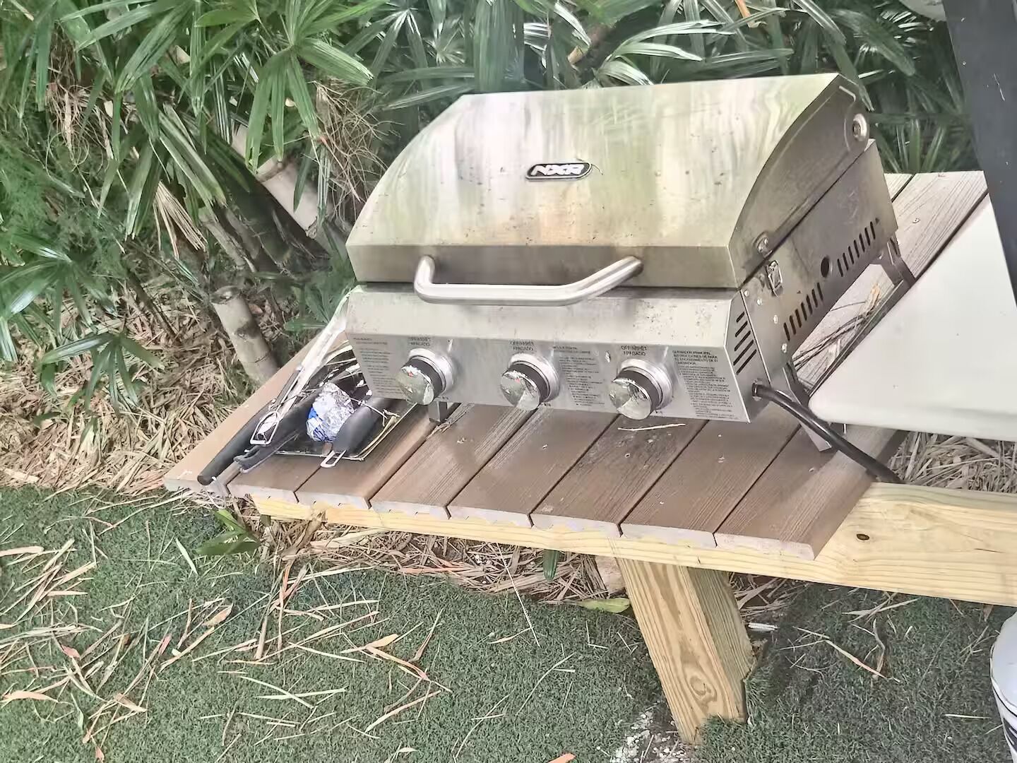 A portable gas grill with four burners and a side burner, placed on a wooden table outdoors.