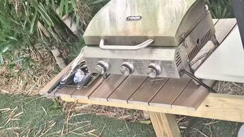 A portable gas grill with four burners and a side burner, placed on a wooden table outdoors.