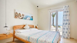 Ein Schlafzimmer mit einem hölzernen Bett, einem Nachttisch, einem Bild einer Küstenlandschaft und Meerblick durch eine Glastür.