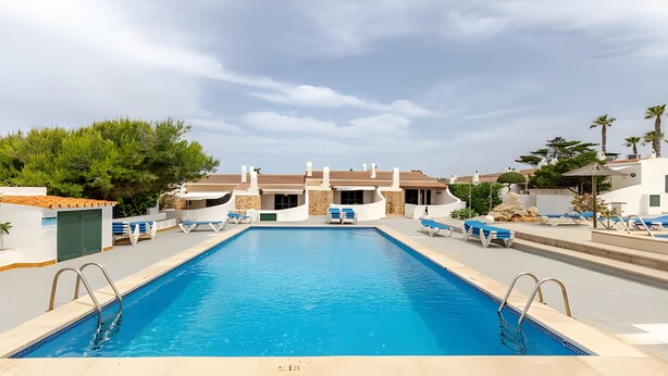 A swimming pool with lounge chairs and a building with a curved roof in the background. - Apartment 'Villa Estrellas 2' with Shared Pool, Private Terrace, and Wi-Fi (Cap d'Artrutx)