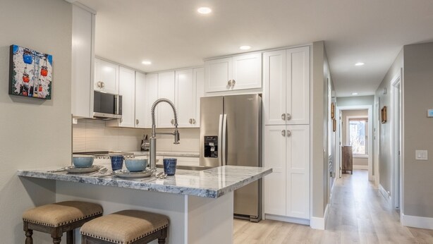 A modern kitchen with white cabinets, a stainless steel refrigerator, a marble countertop, and two bar stools. - Snowcreek IV 559 | Dog Friendly (Mammoth Lakes)