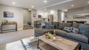 A modern living room with a grey sectional sofa, a wooden coffee table with a bottle and glass, and a kitchen in the background. - Snowcreek IV 559 | Dog Friendly (Mammoth Lakes)