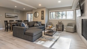 A spacious living room with a grey sectional sofa, a dining area with a table and chairs, and a large window providing natural light. - Snowcreek IV 559 | Dog Friendly (Mammoth Lakes)