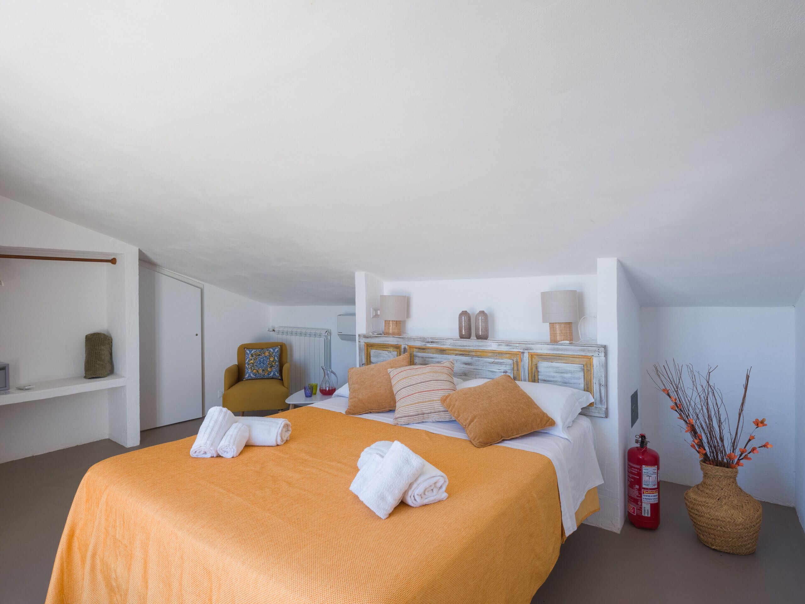 Une chambre d’hôtel avec un lit, une chaise, une étagère, un extincteur et un vase décoré de branches.