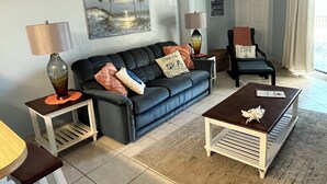 Ein Wohnzimmer mit einem dunkelblauen Sofa, zwei Beistelltischen mit Lampen, einem Couchtisch und einem Palmen-Gemälde an der Wand.