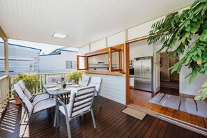 Un espacio exterior cubierto para comer con una mesa y sillas, una terraza de madera y vista a la cocina a través de una puerta de vidrio.