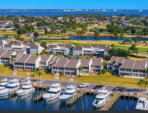 A marina with multiple boats docked, a golf course in the background, and modern buildings with a uniform architectural style. - Harbor-view 1BR with sunsets, full amenities, and family-friendly space. (Panama City Beach)