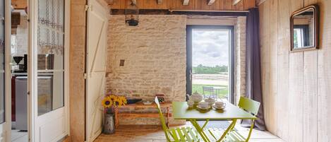 A rustic room with a stone wall, wooden ceiling, a table with chairs, a window with a view, and a door.
