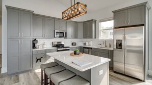 Une cuisine moderne avec des meubles gris, un réfrigérateur en acier inoxydable, un îlot central doté de tabourets et un luminaire suspendu.