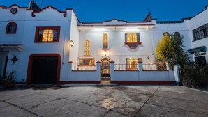 Un edificio blanco con detalles en rojo, una entrada protegida por una reja y varias ventanas con ornamentación de hierro.