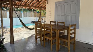A wooden table and chairs under a wooden pergola, with a pool and a white door in the background. - Oceana Suites Vila Cajarana (Tibau do Sul)