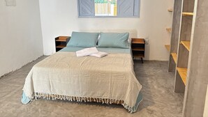 A bedroom with a bed, two pillows, a blanket, a window with blinds, and a wooden shelf. - Oceana Suites Vila Cajarana (Tibau do Sul)