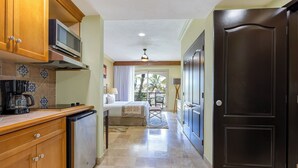 A hotel room with a kitchenette, a bed, a balcony with a view of palm trees, and a dark wooden door. - Cozy 1-bedroom luxe hotel suite in magnificent and fun Cabo San Lucas. (Cabo San Lucas)