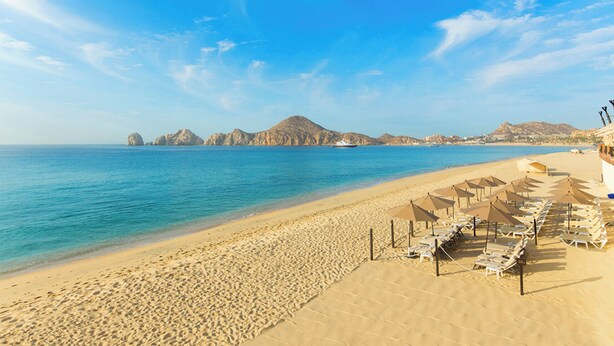 A beach with rows of sun loungers and umbrellas, a sandy shore, and distant mountains. - Cozy 1-bedroom luxe hotel suite in magnificent and fun Cabo San Lucas. (Cabo San Lucas)
