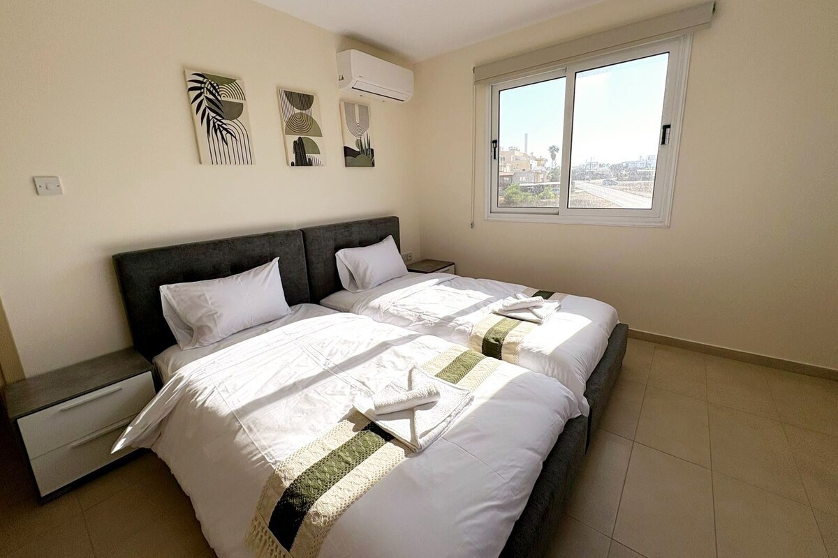A bedroom with two beds, a nightstand, and a window with a city view.