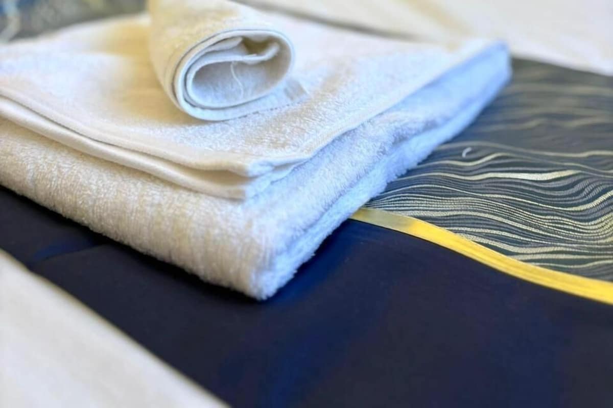 A neatly made bed with a rolled white towel and a folded white towel on a bed with a dark blue and yellow patterned bedspread.