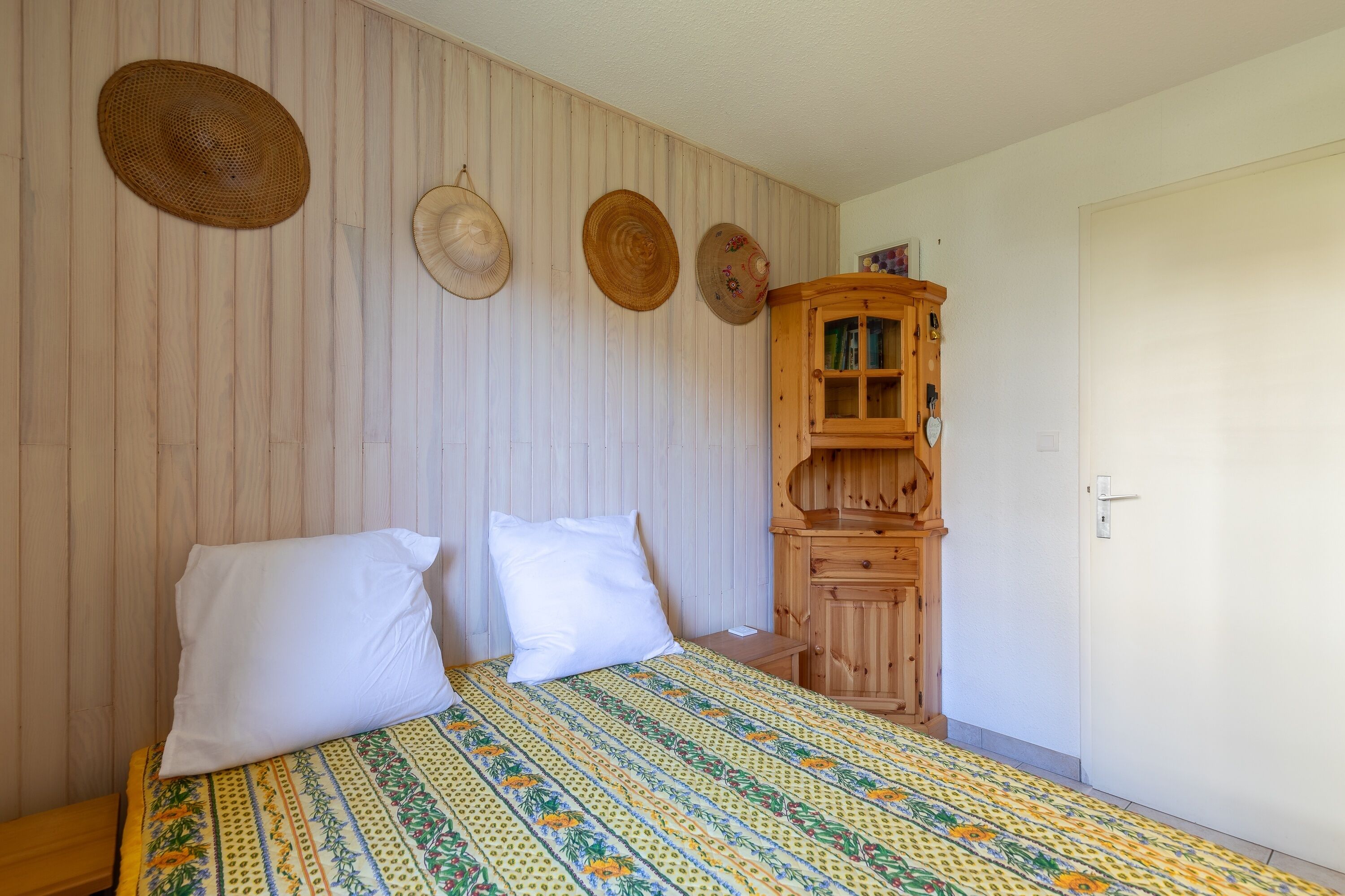 Une chambre à coucher avec un lit, deux oreillers, une armoire en bois et trois chapeaux accrochés au mur.