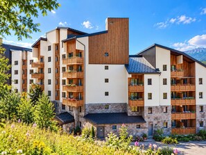 A multi-story hotel with balconies, stone facade, and a mountainous backdrop. - Les Bergers - Proche Station de Ski (Huez)