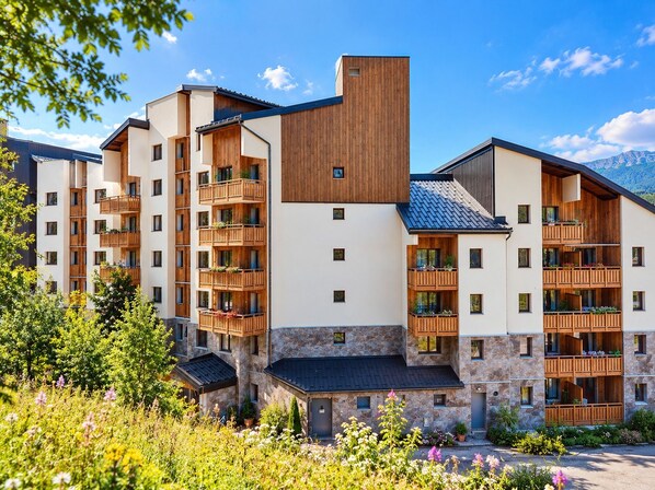 A multi-story hotel with balconies, stone facade, and a mountainous backdrop. - Les Bergers - Proche Station de Ski (Huez)