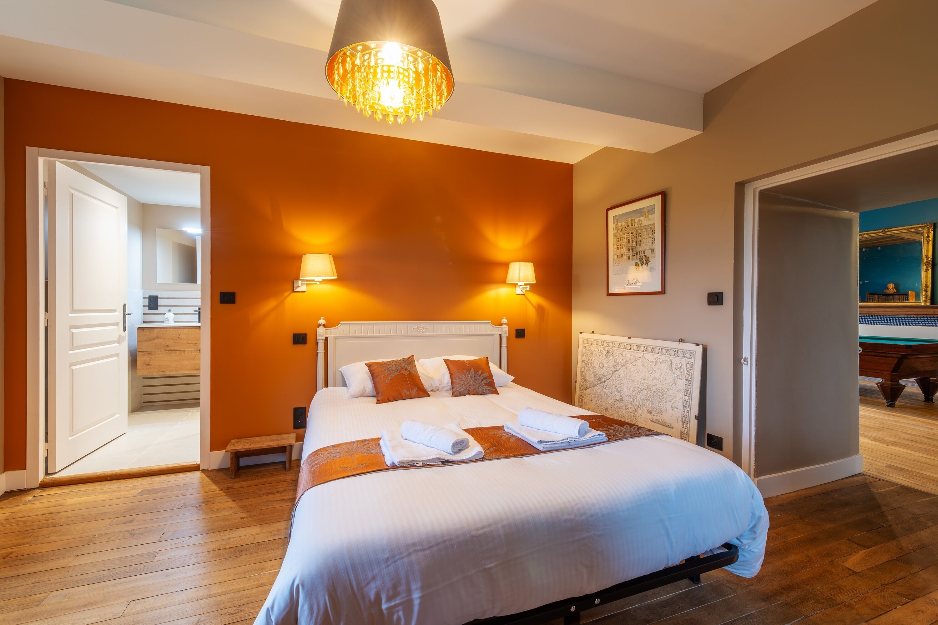 A hotel room with a large bed, two bedside lamps, a chandelier, and a painting on the wall.