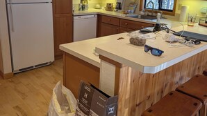 Une cuisine avec des meubles en bois, un réfrigérateur blanc, un four à micro-ondes et un lave-vaisselle. Une fenêtre offre une vue sur les arbres.
