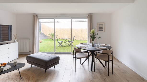 A modern living room with a dining area, a grey sofa, a TV, and a view of a garden through large sliding doors. - Couleur Sable - 1km de la Plage (Dinard)