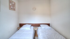 Ein kleiner, sauberer Raum mit zwei Einzelbetten, einem hölzernen Kopfteil, einem kleinen Hocker und einer Deckenleuchte.
