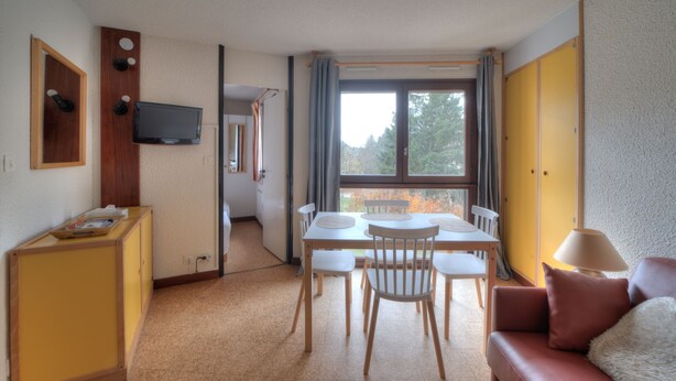 Ein kompakter Wohnbereich mit Essplatz, einer Couch, einem an der Wand montierten Fernseher und einem Fenster mit Blick auf Bäume.