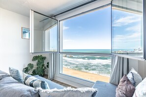 A room with a large window overlooking a beach, a sofa with cushions, and a potted plant. - Luna - Studio vue imprenable sur la mer (Biarritz)