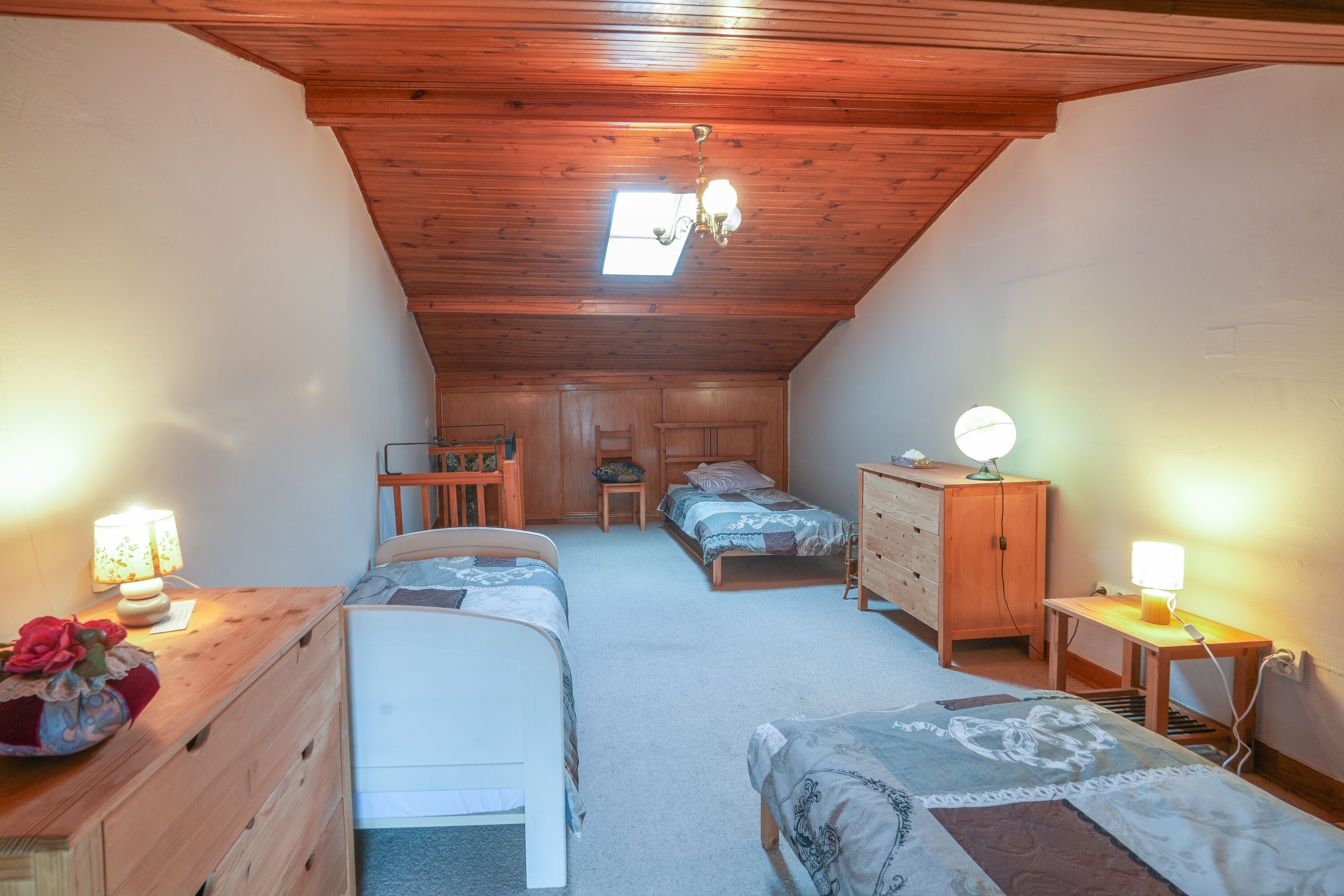 Une chambre mansardée chaleureuse, avec deux lits, une commode en bois, un bureau avec une lampe et une fenêtre de toit.