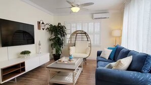 Un salon moderne avec un téléviseur à écran plat, un canapé blanc, un canapé bleu, une table basse, un fauteuil en osier et une plante en pot.