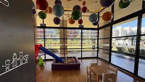 A children's play area with a slide and ball pit, large windows with a view of buildings, and a sign indicating the exit. - Noroeste design comfort and charm (Brasília)