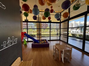 A children's play area with a slide and ball pit, large windows with a view of buildings, and a sign indicating the exit. - Noroeste design comfort and charm (Brasília)