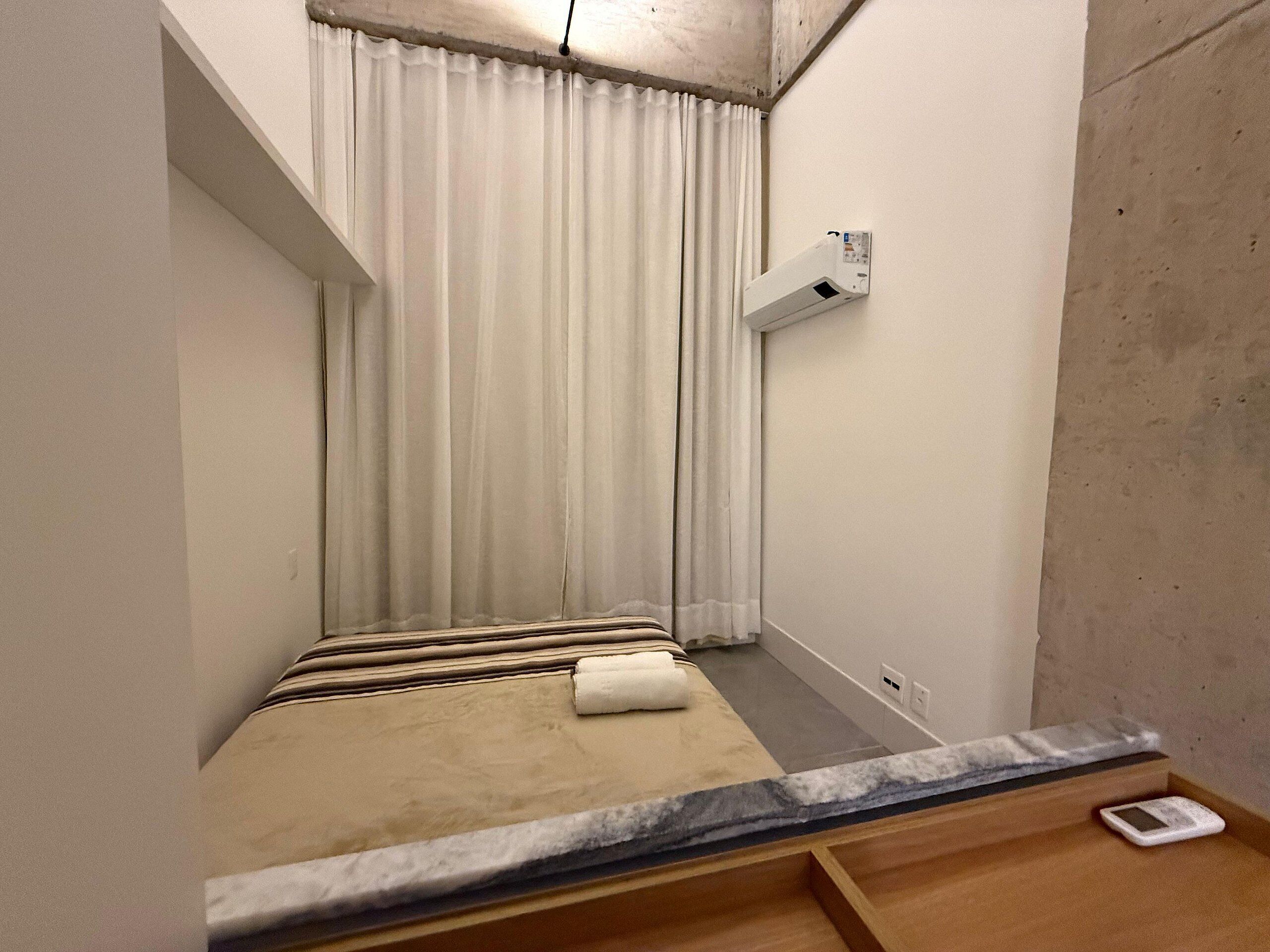 A small, narrow room with a bed, a white air conditioner, and a window with white curtains.