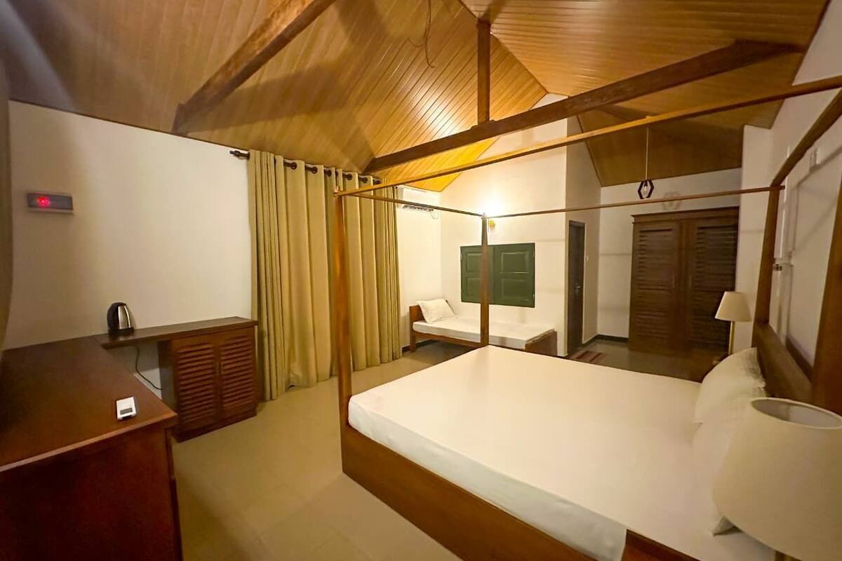 A hotel room with a wooden ceiling, a bed, a bedside table with a lamp, a small desk with a kettle, and a window with curtains.