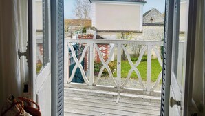 Uitzicht door een deur met een witte metalen balustrade, leidend naar een balkon met een heldere lucht en een gebouw met een pannendak op de achtergrond.