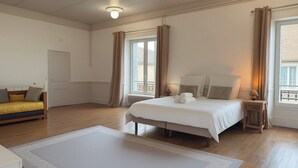 Een slaapkamer met een bed, nachtkastje, lamp, raam met gordijnen, radiator en een bank met kussens.