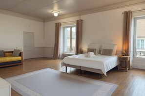 A bedroom with a bed, a bedside table, a lamp, a window with curtains, a radiator, and a sofa with cushions.
