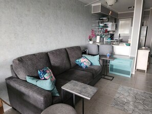 A modern living room with a grey sofa, teal cushions, a small side table, and a kitchen area with white cabinets and a teal countertop. - Coastal Paradise 2B/2B with the beach atmosphere you want at Hilton Head! (Hilton Head Island)