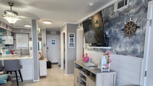 Moderni keittiö, jossa on sisäänrakennettu mikroaaltouuni, ruokailutila, jossa on harmaa sohva ja pieni pöytä, seinälle asennettu televisio sekä koristeellinen laivan ruori.