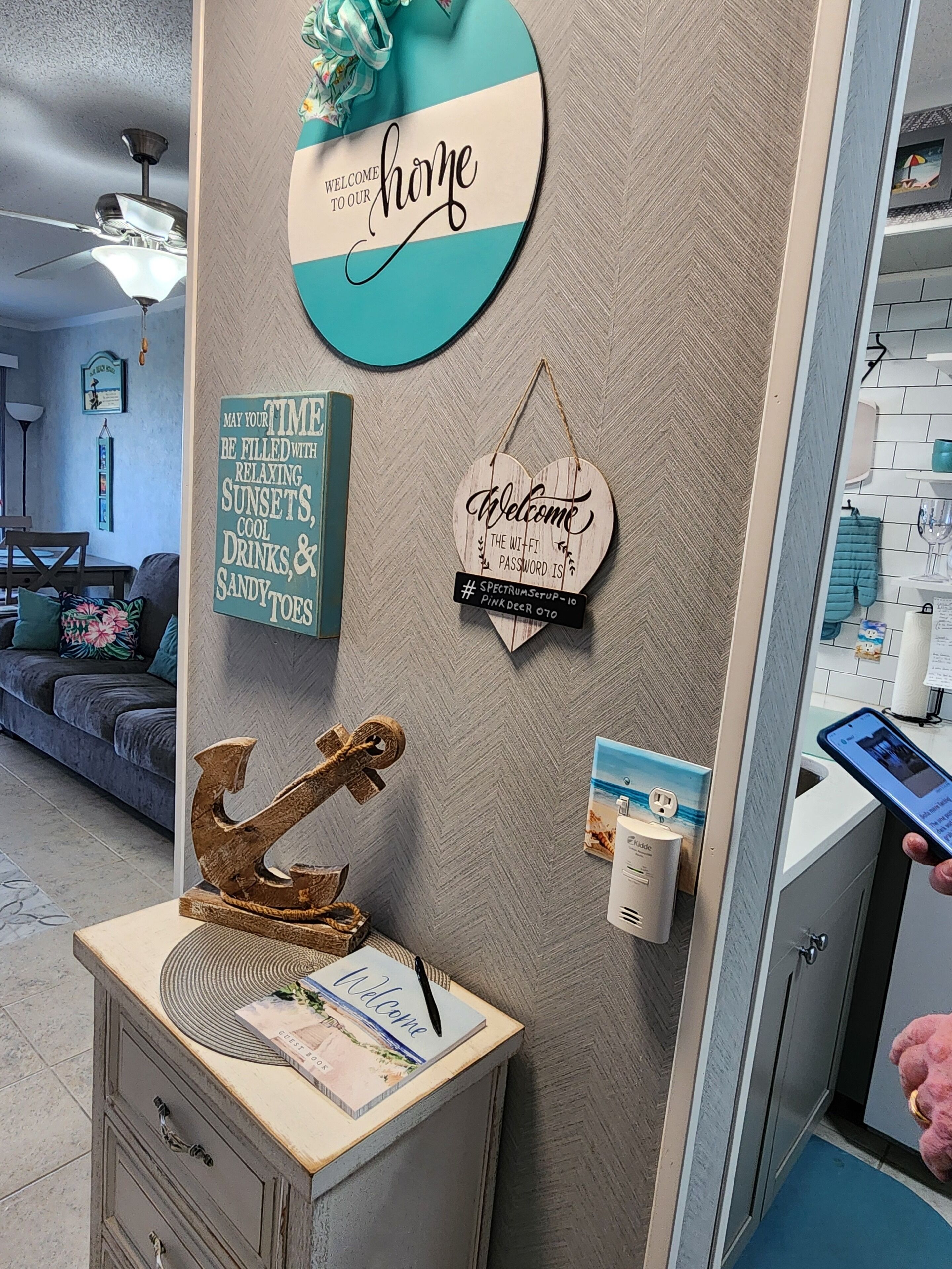 A room with a wall decorated with various signs, including a 'Welcome home' sign, a sign about relaxing time, and a Wi-Fi password sign. There is a desk with a book and a pen, and a small decorative anchor.