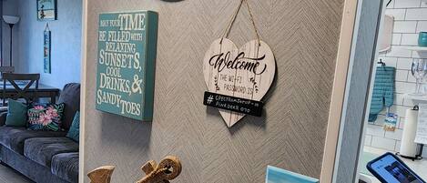 A room with a wall decorated with various signs, including a 'Welcome home' sign, a sign about relaxing time, and a Wi-Fi password sign. There is a desk with a book and a pen, and a small decorative anchor.