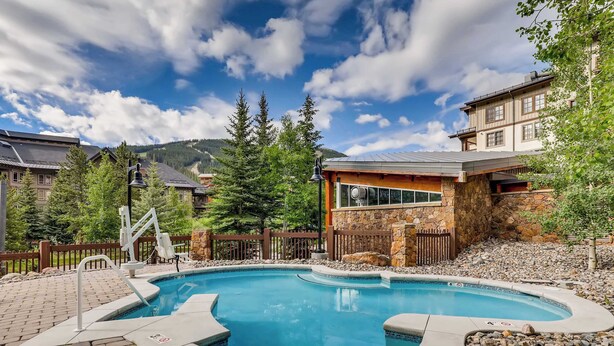 Une piscine bordée d’un mur en pierre et dotée d’une terrasse en bois, entourée d’arbres et offrant une vue sur la montagne.